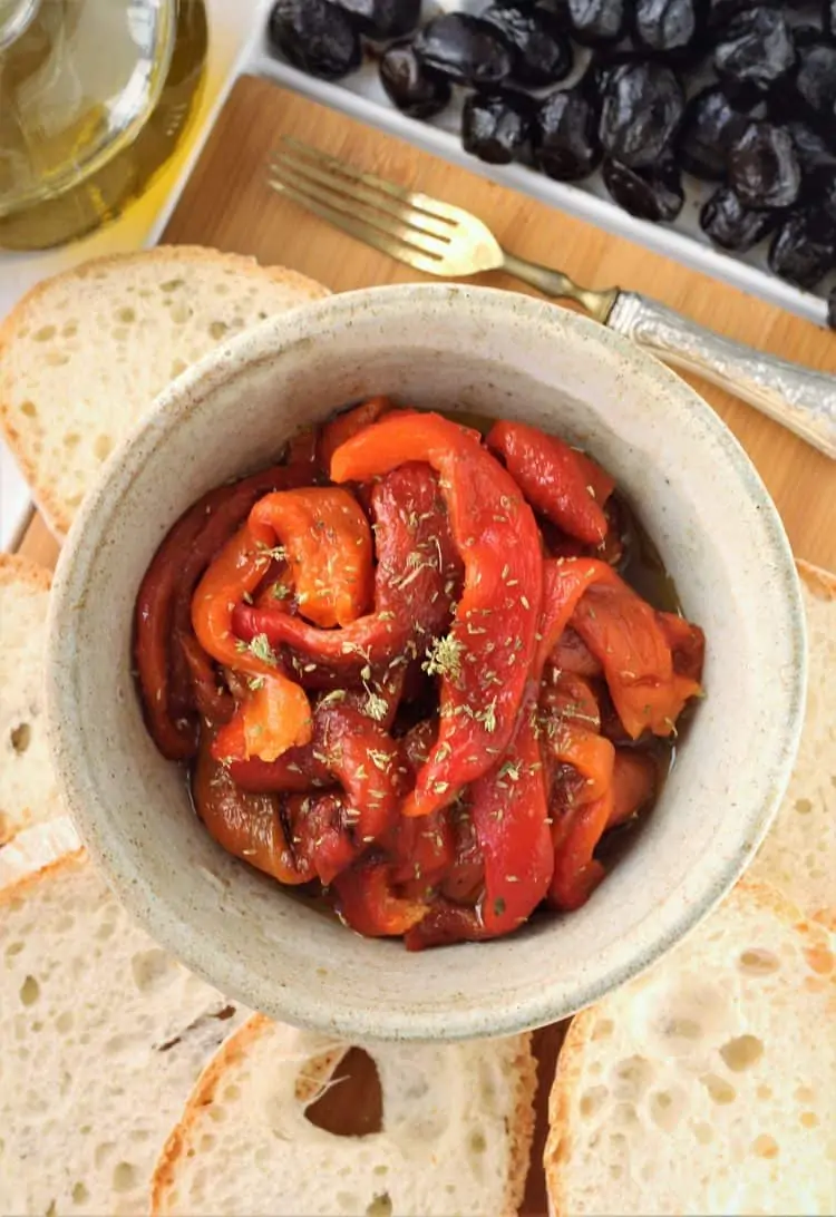 bowl of roasted red bell pepper strips surrounded by bread slices, olives and olive oil 