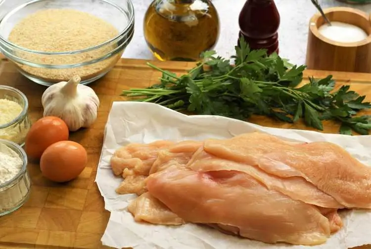 raw chicken cutlets on wood board with eggs, garlic, bowl of breadcrumbs, olive oil, salt and pepper