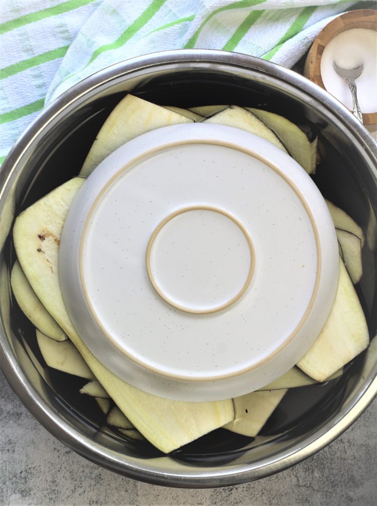 large bowl filled with eggplant slices in water with plate covering them