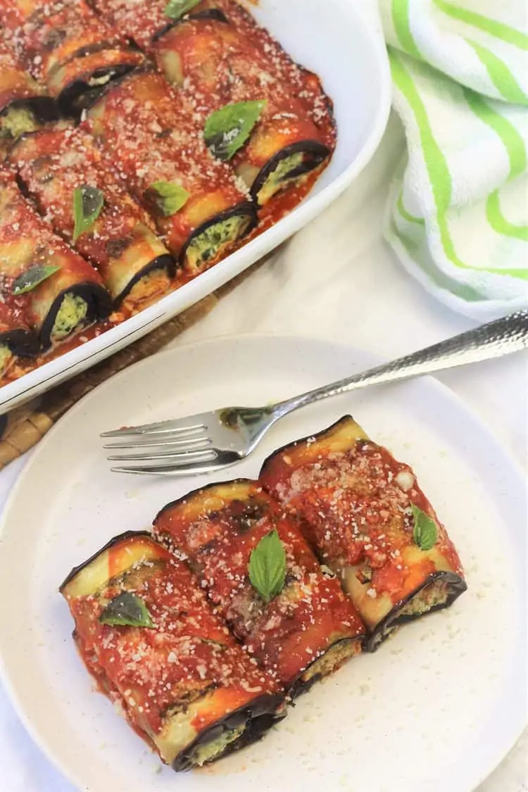 eggplant involtini on white plate with fork and baking dish filled with rolls behind it
