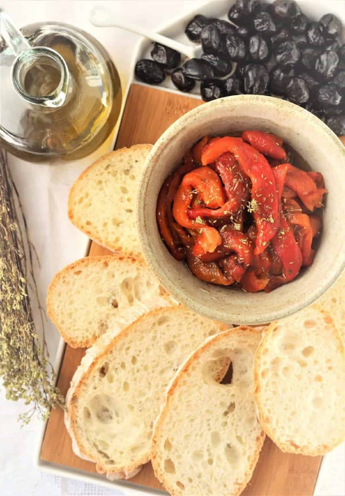 Marinated roasted red bell peppers in bowl surrounded by sliced bread, olives and olive oil.
