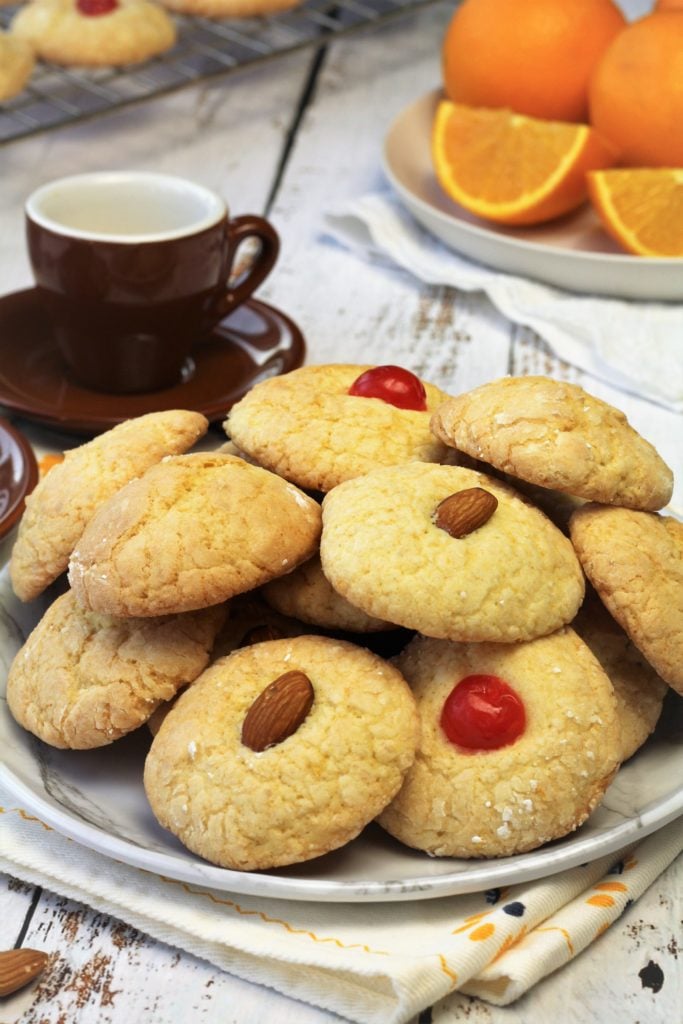 orange juice cookies with almonds and maraschino cherries piled on plate with espresso cups and oranges in background