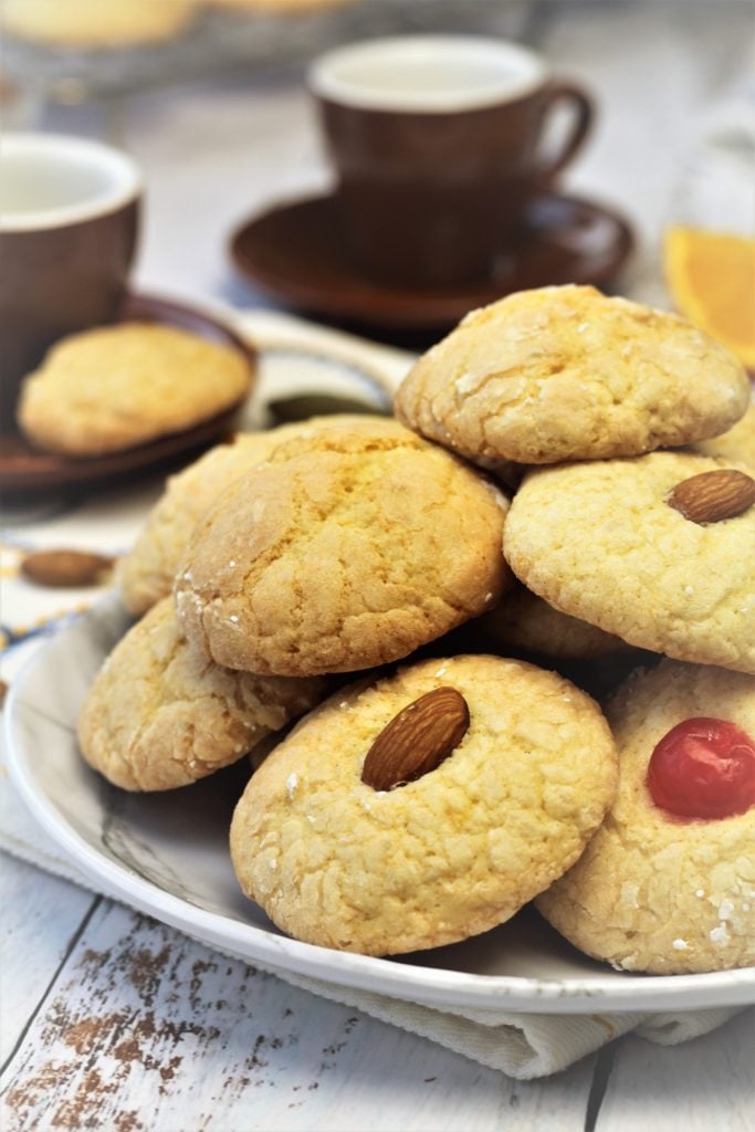 orange juice cookies topped with almonds and maraschino cherries on white plates with espresso cups in background