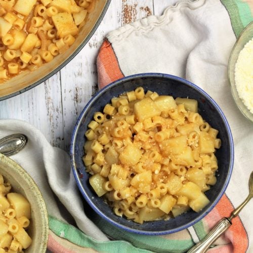 bowl of pasta with potatoes surrounded by pot, spoon and cheese bowl