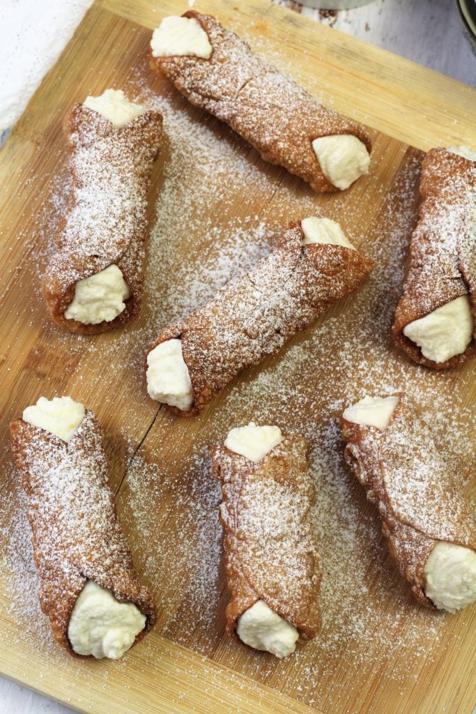 cannoli shells on wood board filled with ricotta and dusted with powdered sugar