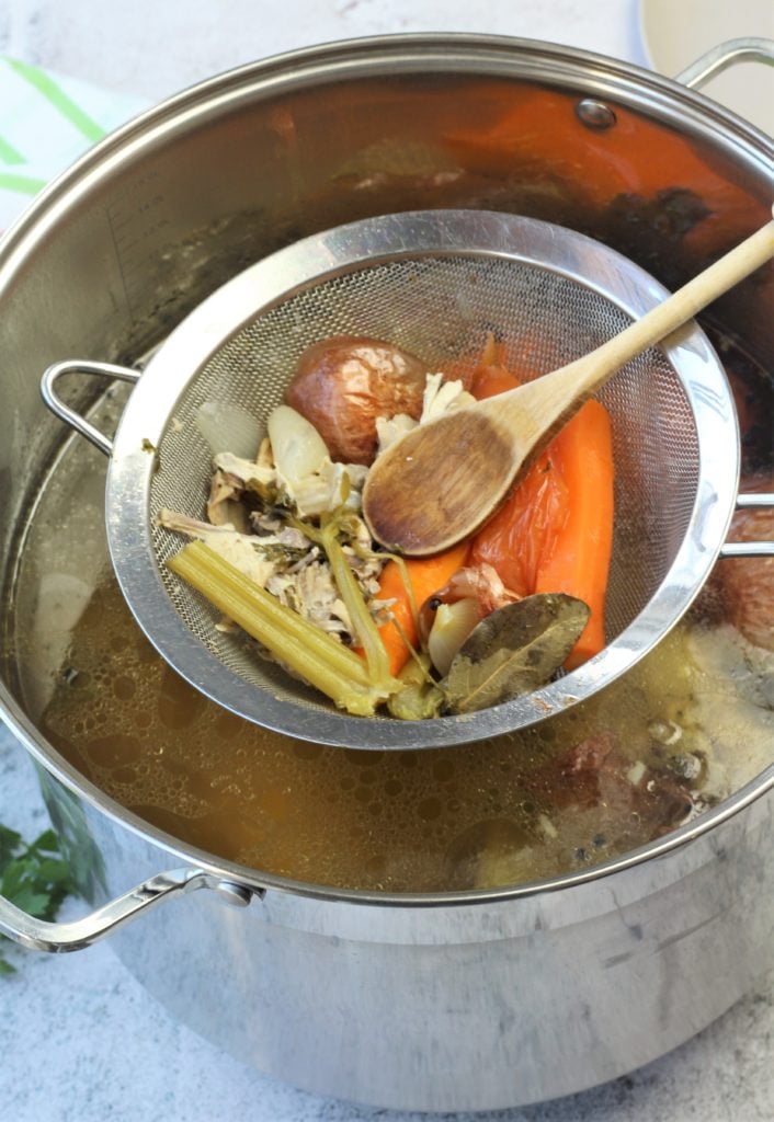 vegetable and chicken pieces in fine mesh sieve with wood spoon held over large stock pot