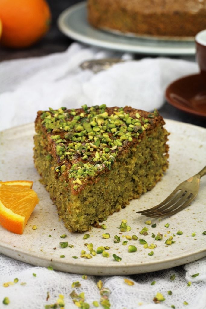 Pistachio cake slice on white plate with orange slices and fork.