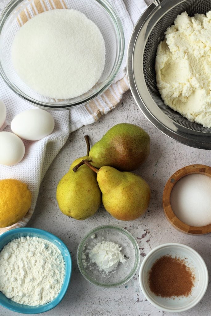 Bowls with flour, sugar, ricotta, cinnamon alongside eggs, pears and a lemon.