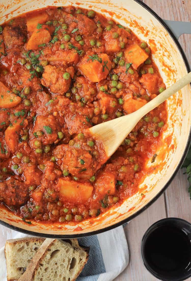 Large skillet with wooden spoon stirring meatball stew with potatoes and peas.