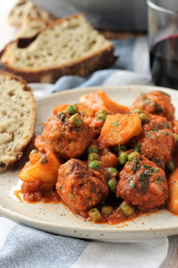 Plated meatballs with potaoes and peas in tomato sauce with sliced bread.