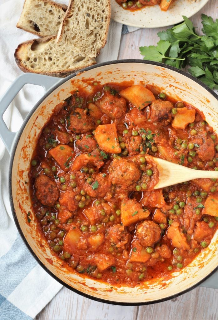 Skillet filled with meatball stew with potatoes and peas and wood spoon stirring. 