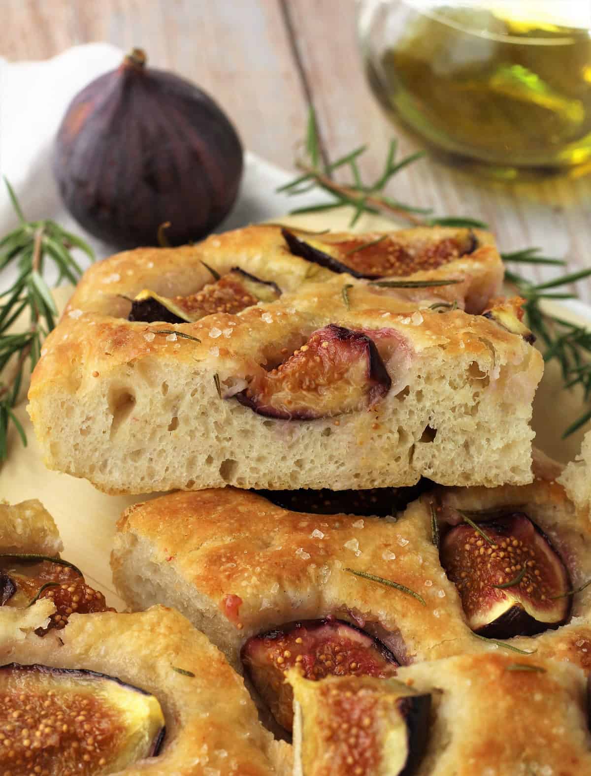 No knead fig and rosemary focaccia squares piled next to fig, olive oil bottle and rosemary figs.