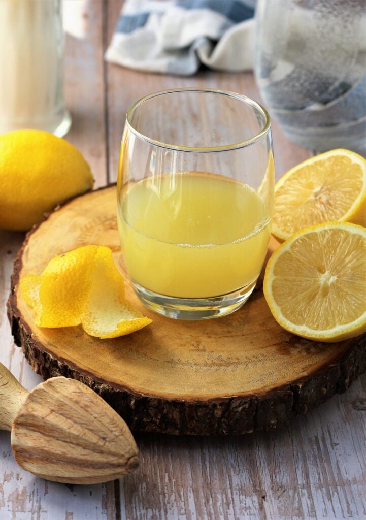 Glass filled with lemon juice with cut lemons on wood board.