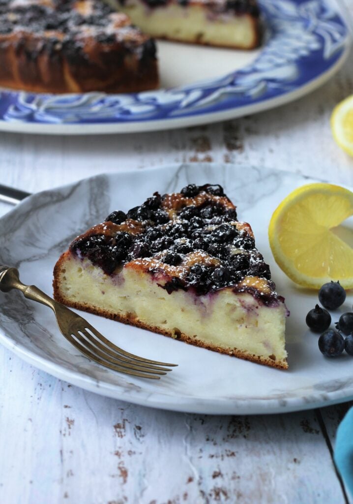 Plated slice of ricotta cake with blueberries and lemon.