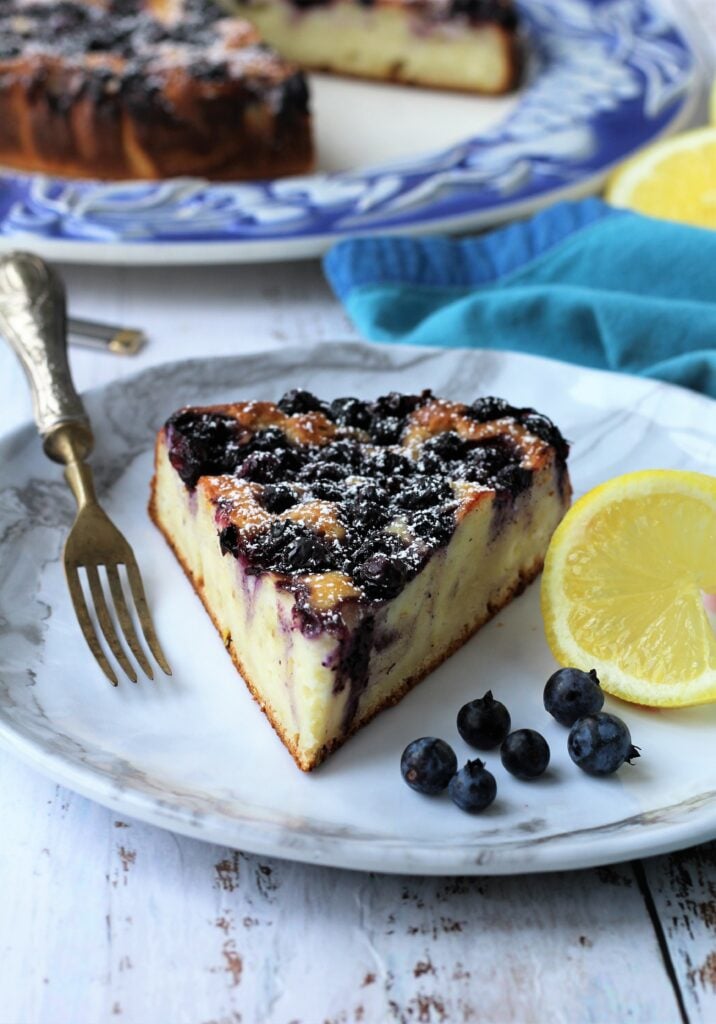 Slice of blueberry ricotta cake on plate with blueberries and lemon wedge.