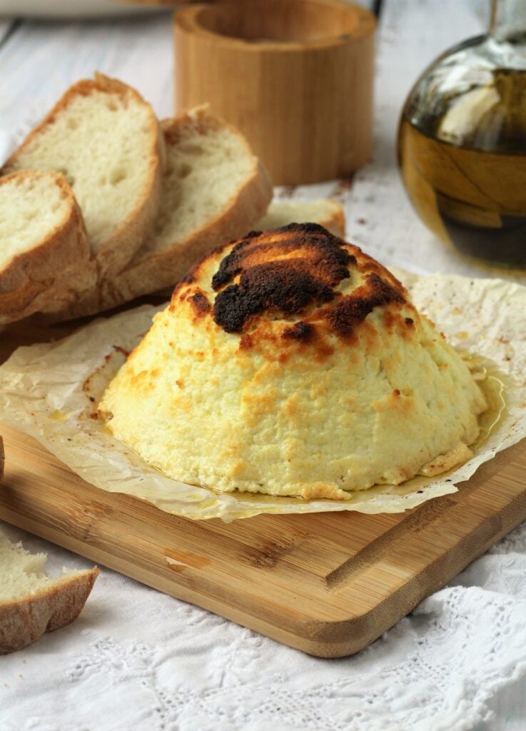 Whole baked Sicilian ricotta on wood board with bread slices.