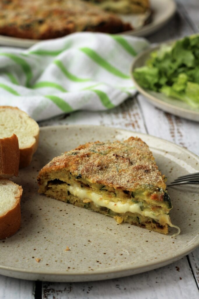 Wedge of cheese filled zucchini frittata on plate with bread and salad.