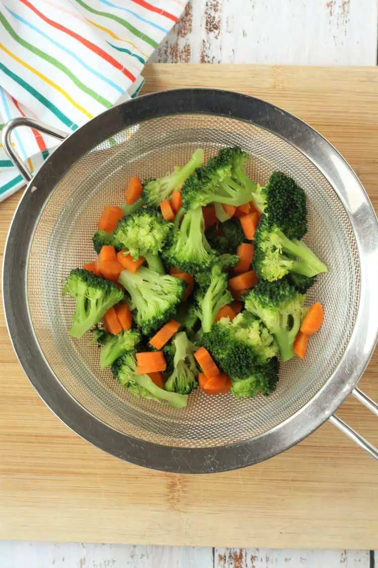 Broccoli florets and carrots in sieve over bowl.