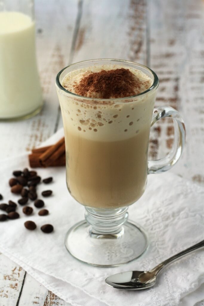Glass cup with iced cappuccino with spoon, coffee beans and bottle of milk on side.