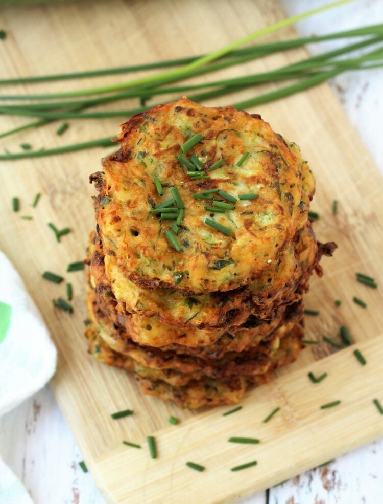 Zucchini fritters piled on wood board with chives on side.