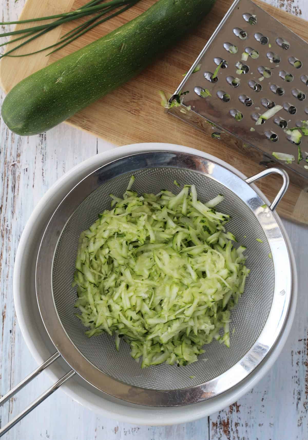 Shredded zucchini in fine mesh sieve over bowl next to box grater.