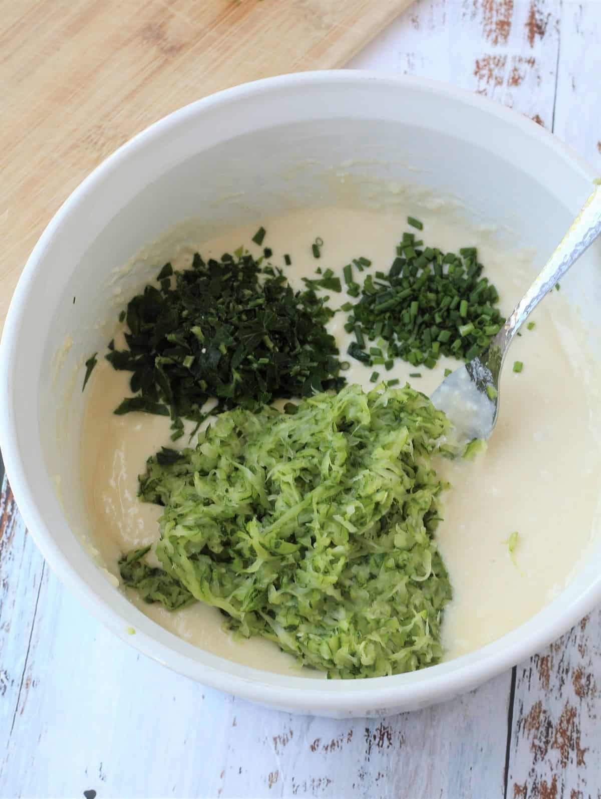 Shredded zucchini and herbs added to bowl with batter for zucchini fritters.