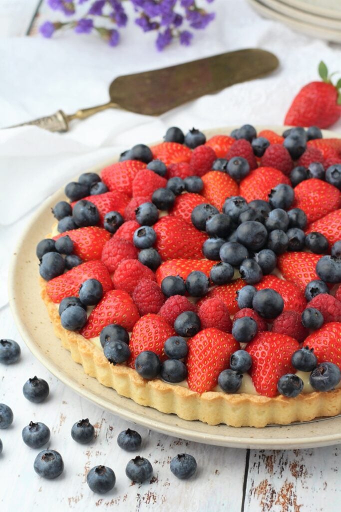 Fruit tart with custard filling on plate with blueberries around it and spatula.
