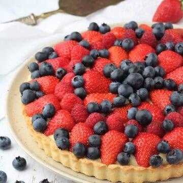 Italian fruit tart with pastry cream filling on plate surrounded by blueberries.