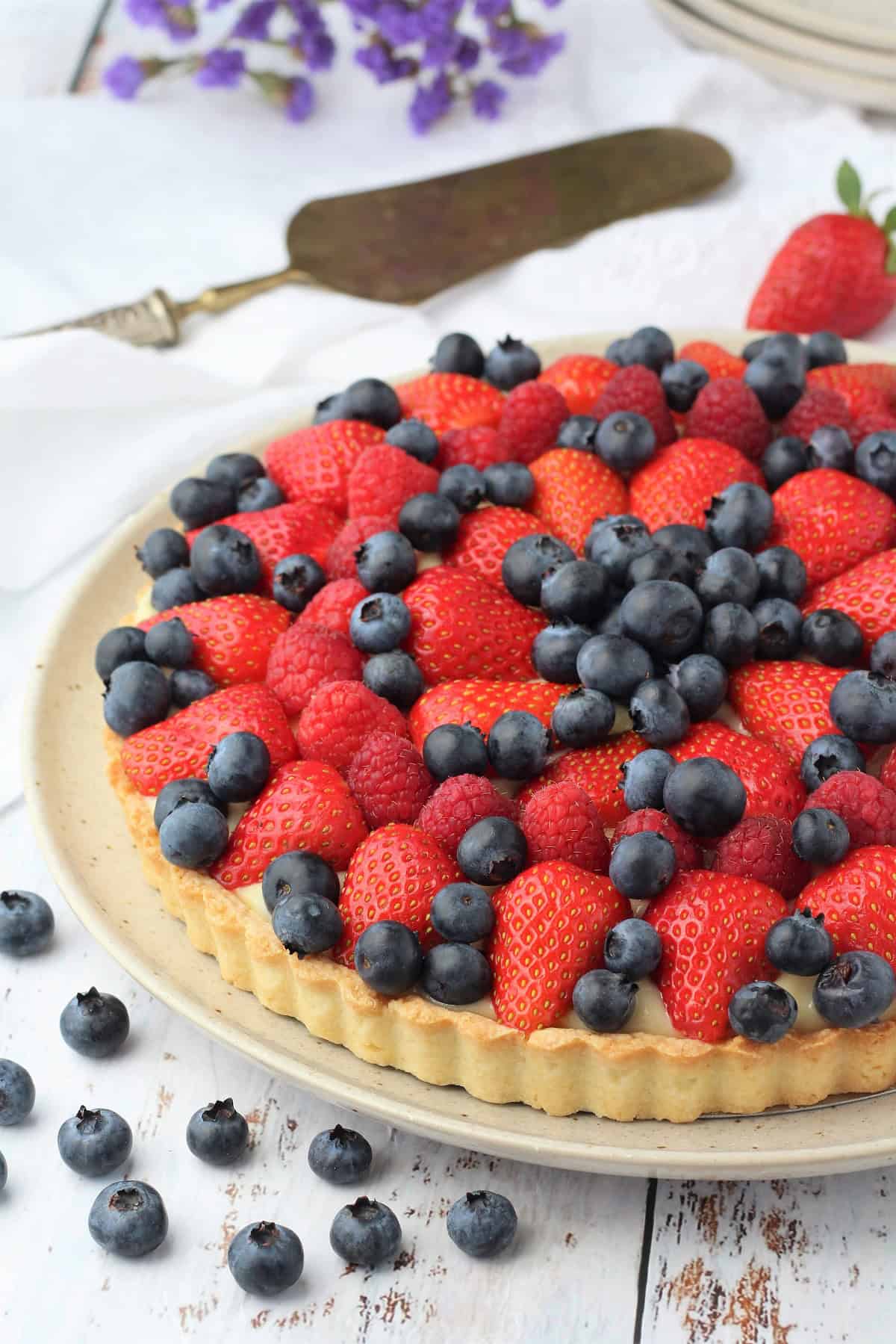 Italian fruit tart with pastry cream filling on plate surrounded by blueberries.