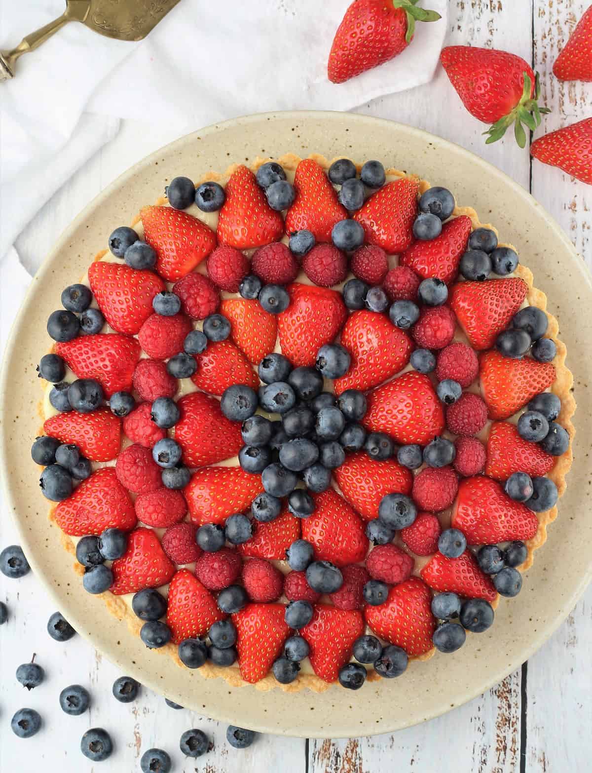 Italian fruit tart with pastry cream topped with berries on round plate surrounded by fruit pieces.