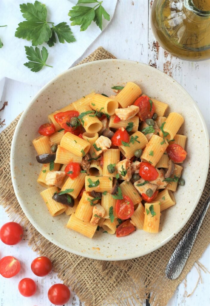 Bowl of swordfish pasta with cherry tomatoes, capers and olives with tomatoes, olive oil and parsley on side.