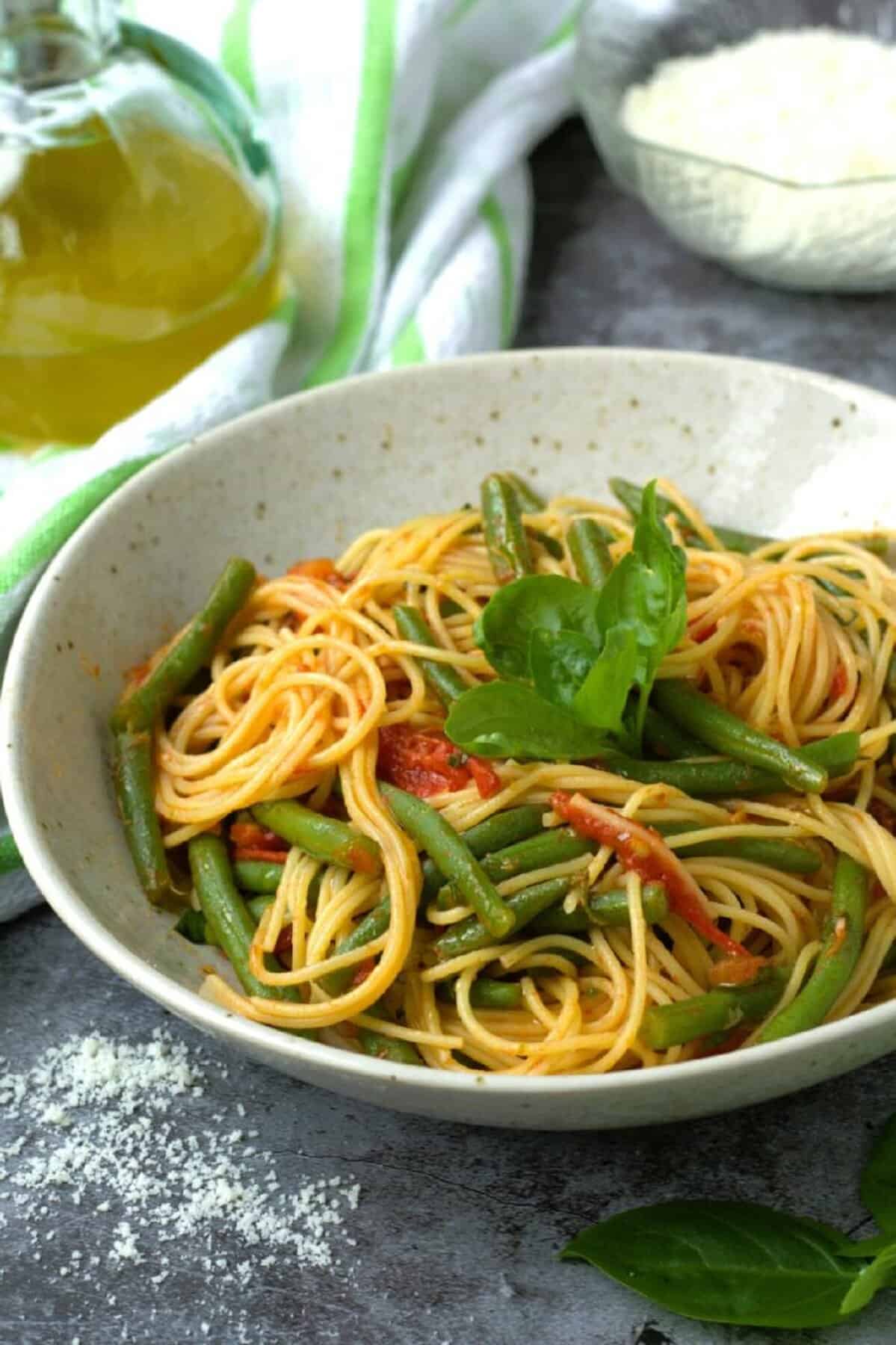 Spaghetti with green beans and tomato sauce in place topped with basil and bowl of Parmigiano cheese next to it.