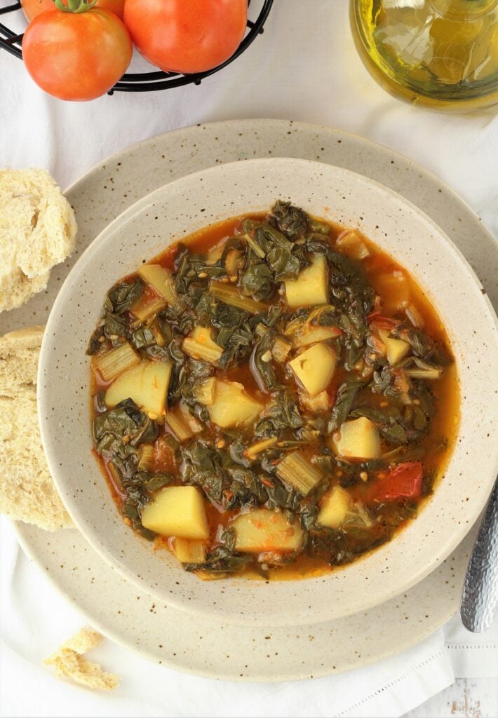 Bowl with swiss chard with potatoes and tomatoes next to bread slices, tomatoes and olive oil.