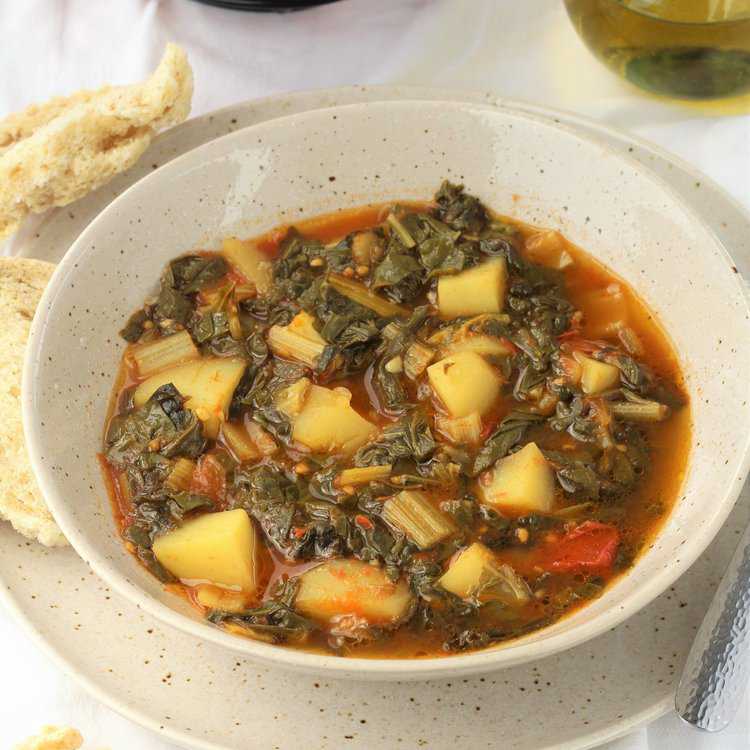 Bowl of swiss chard with potatoes and tomatoes with bread on side.