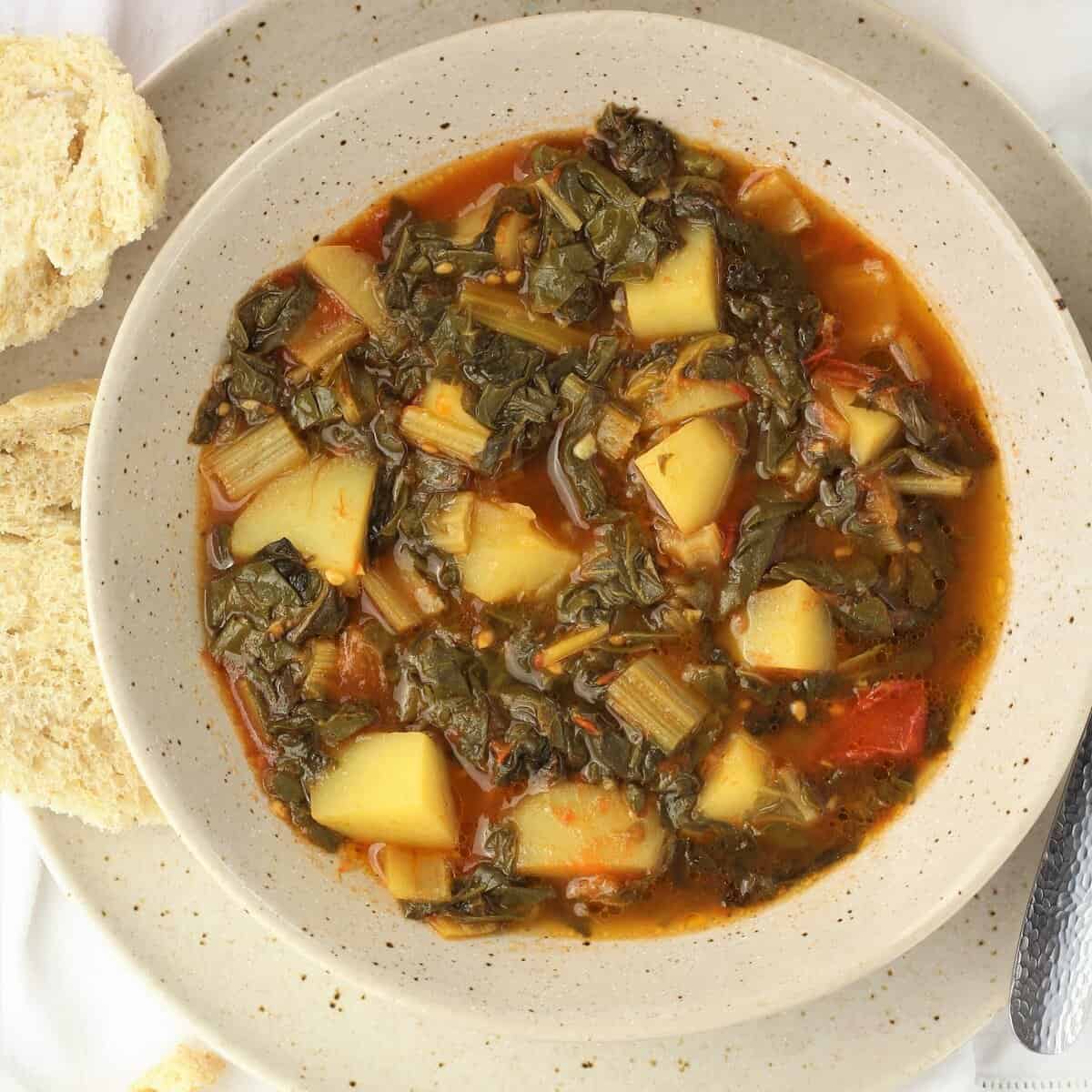 Stewed swiss chard recipe with potatoes and tomatoes in bowl with bread slices next to it.