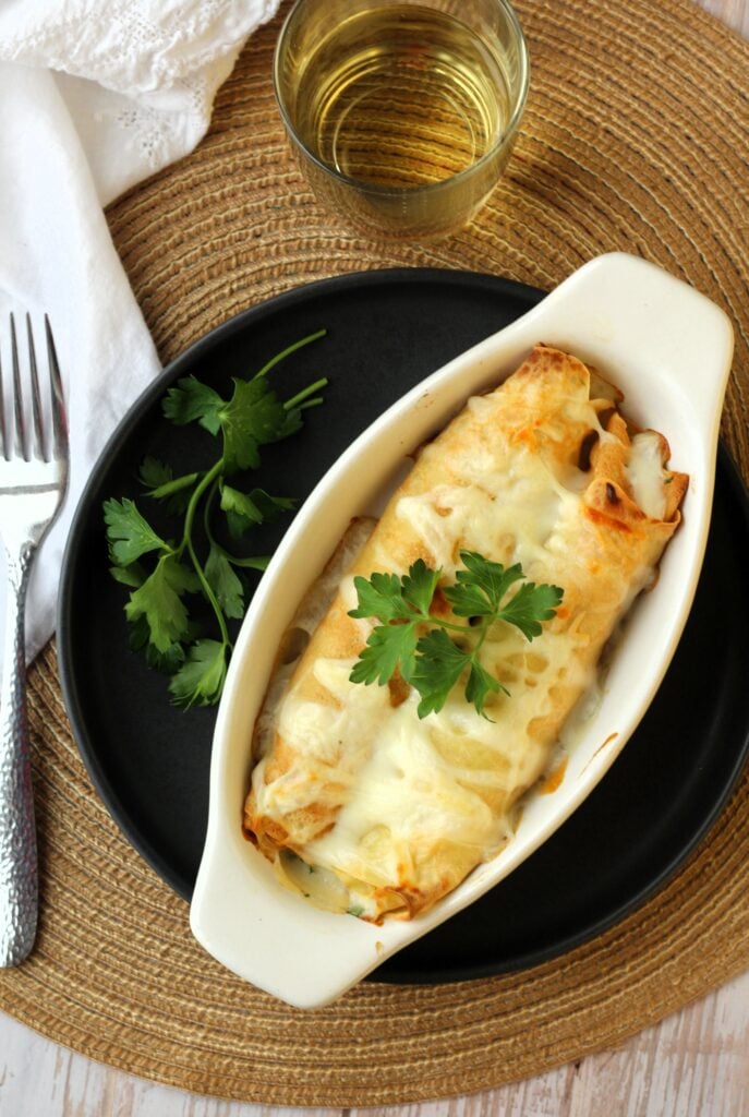 Seafood crepes topped with béchamel sauce in baking dish topped with parsley and wine glass on side.