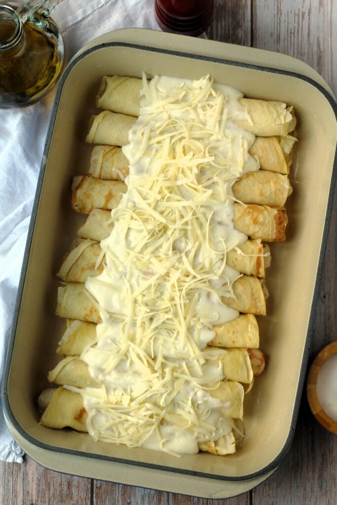 Seafood filled crepes topped with béchamel sauce and cheese in baking dish.