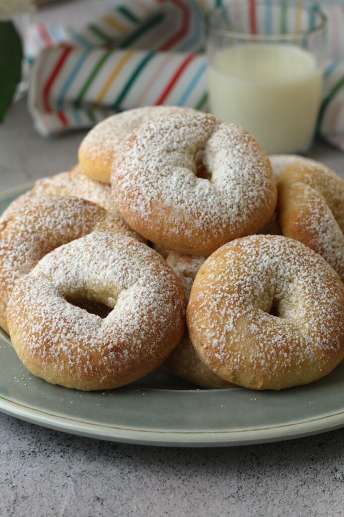 Piled ricotta ring cookies on blue plate with glass of milk behind.