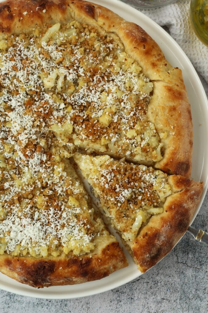 Spatula holding one wedge of cauliflower pizza from plate with whole pizza.