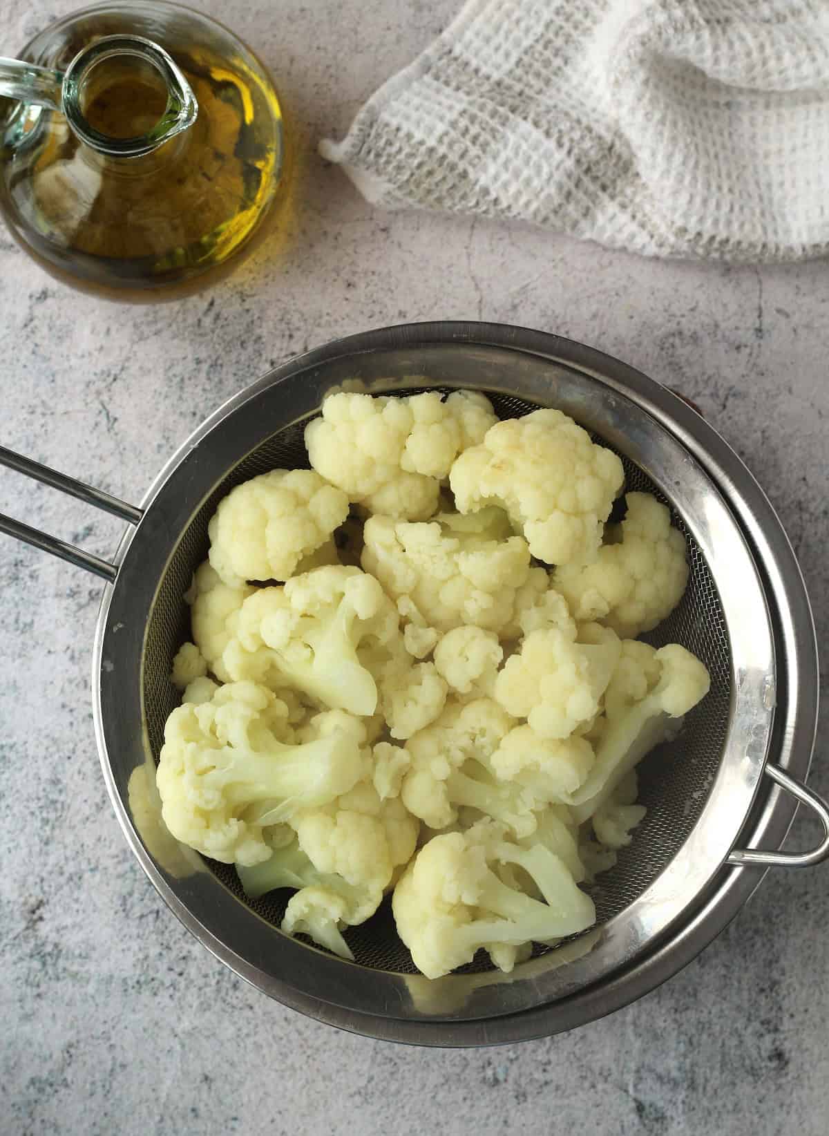Boiled cauliflower in sieve over bowl next to olive oil bottle.