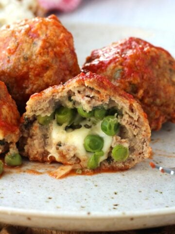 Stuffed meatballs with peas and mozzarella in tomato sauce cut open on plate surrounded by more meatballs.