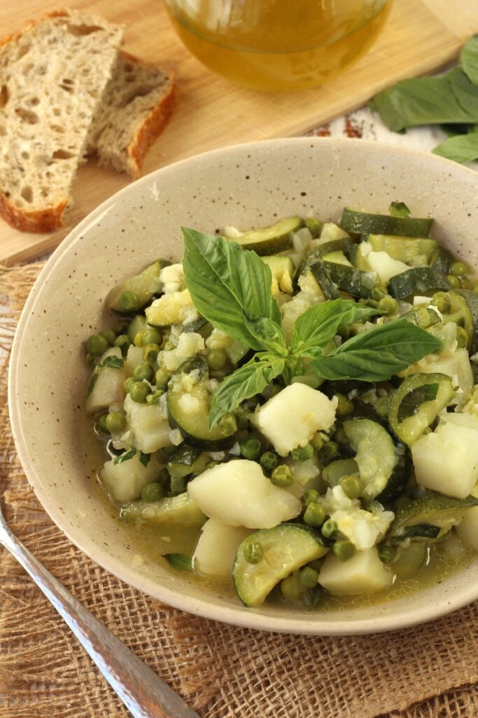 Bowl of zucchini and potatoes with peas and basil along side spoon, bread slices and olive oil.