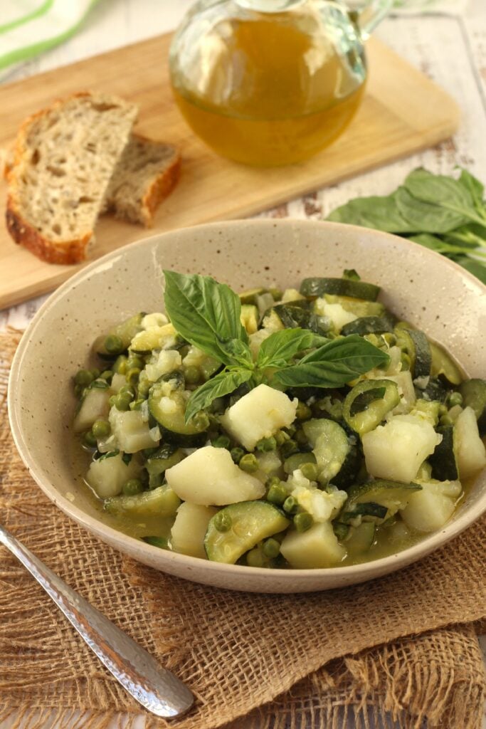 Bowl of stewed zucchini, potatoes and peas with basil along bread slices and bottle of oil.