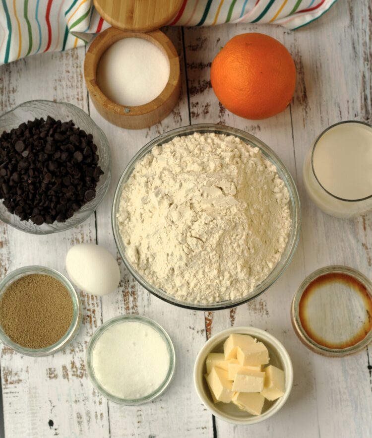 Bowls with flour, butter, sugar, salt, yeast, vanilla, chocolate chips, an egg and an orange.