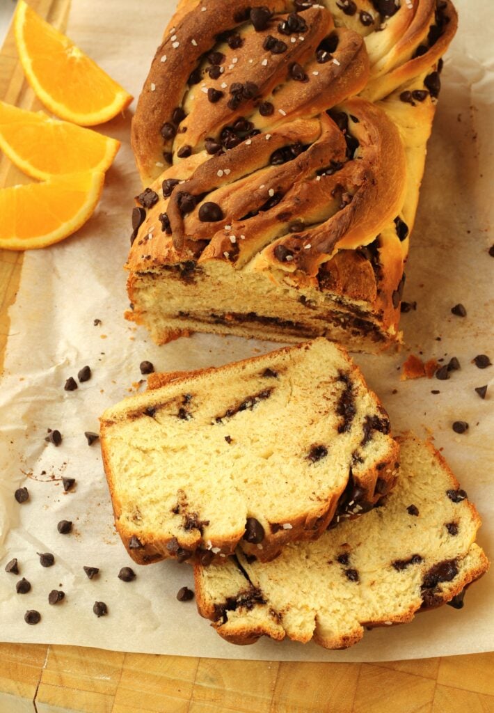 Loaf of sliced chocolate chip twisted brioche bread on wood board with chocolate chips and orange slices.
