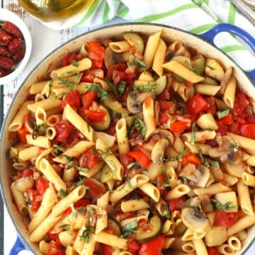 Large skillet filled with vegetable pasta in tomato sauce with olive oil bottle and utensils on side.