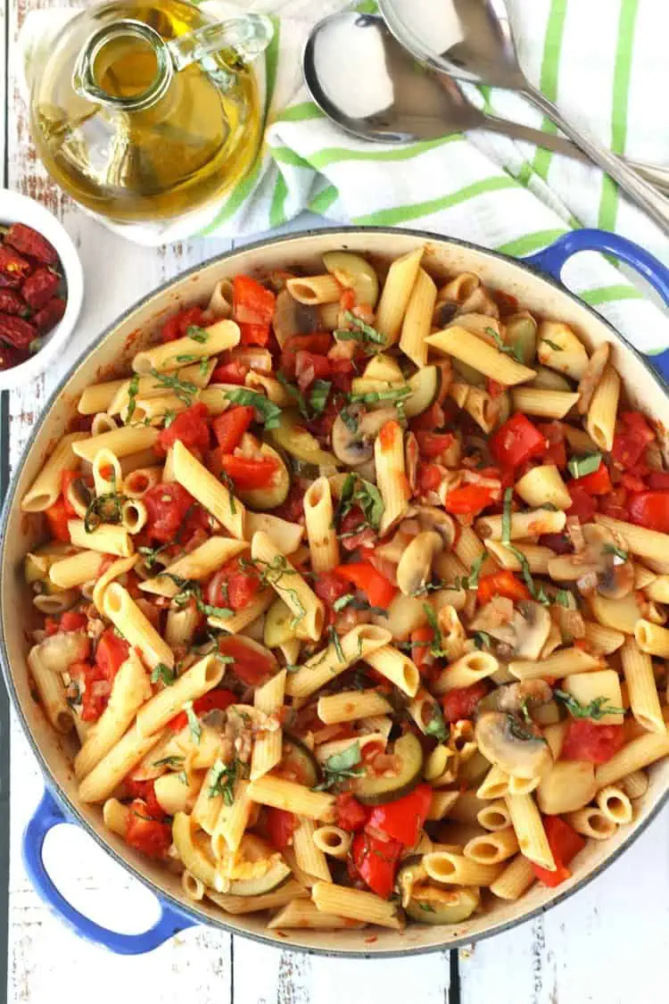 Large skillet filled with vegetable pasta in tomato sauce with olive oil bottle and utensils on side.
