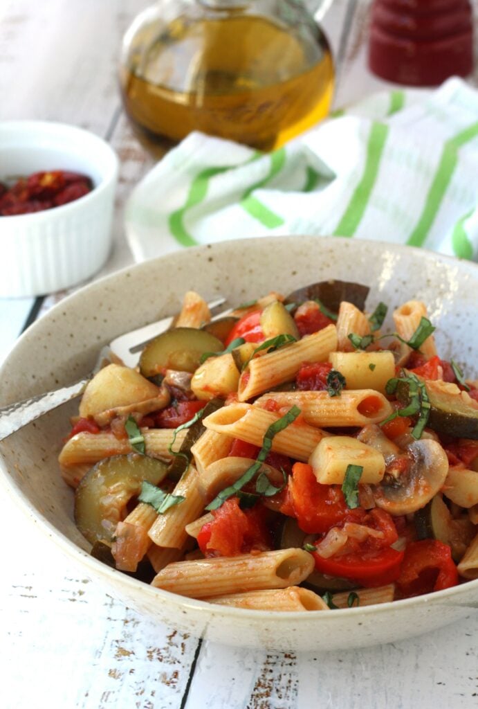 Bowl of penne with vegetables and tomato sauce with olive oil bottle behind it.