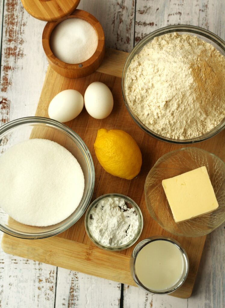 Bowl of flour, sugar, baking powder, salt, butter, eggs, lemon and a glass of milk.