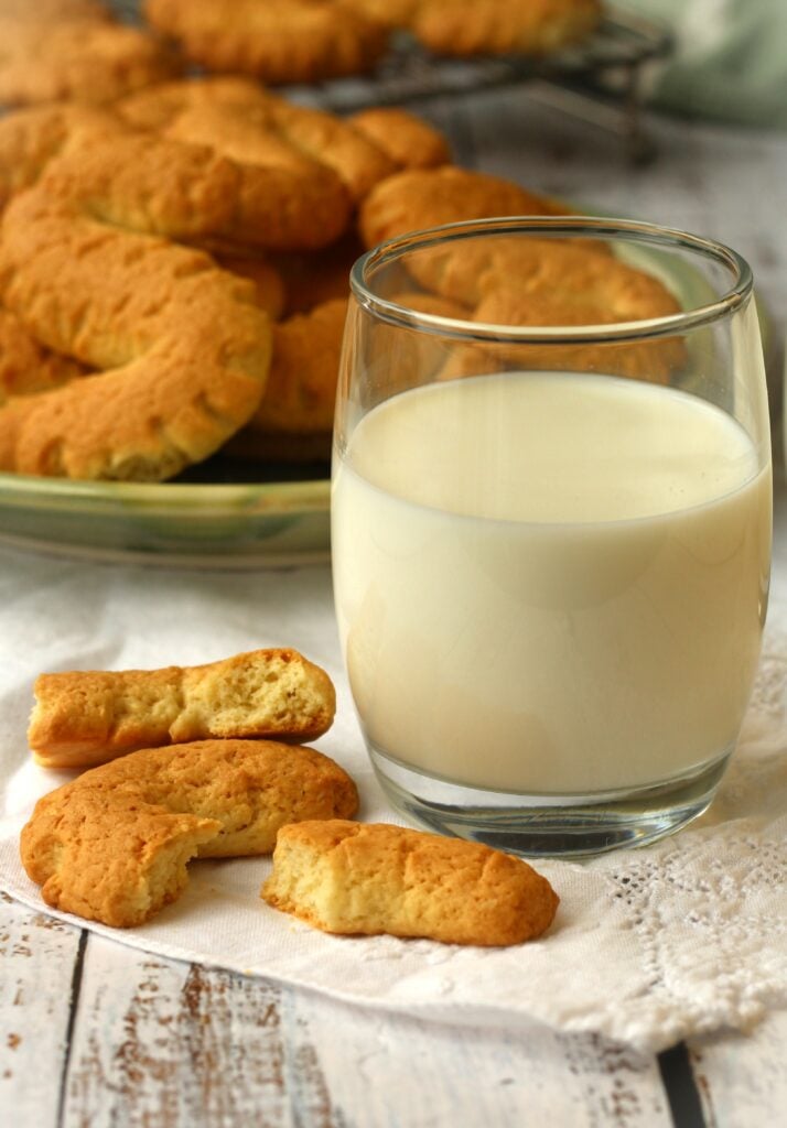 S cookie cut into pieces alongside glass of milk and plate full of cookies behind it.